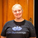 Smiling woman wearing a black On Site Massage t-shirt against a wooden background.